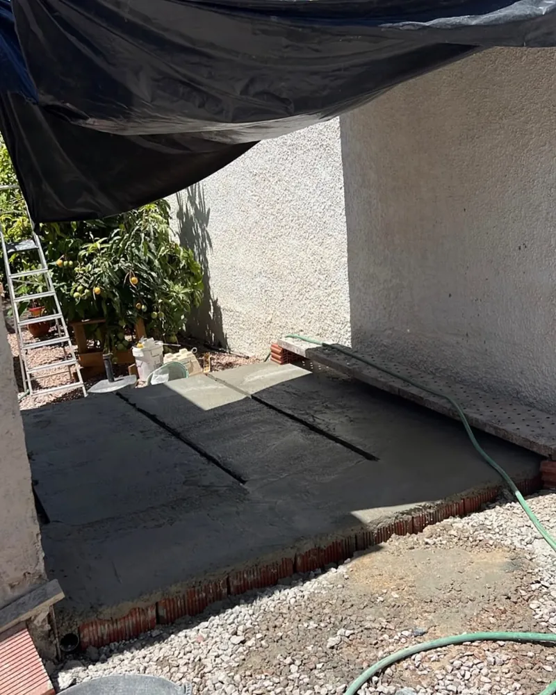 Solera de hormigón recién echada bajo un toldo negro, junto a la pared de la casa